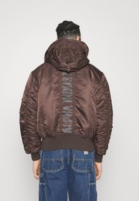 Osoba stojící zády, oblečená v hnědém péřovém kabátu s nápisem "Alpha Industries" na zadní straně a modrých cargo džínách.