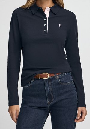 Vrouw draagt een marineblauw poloshirt met lange mouwen, roze biezen en een klein logo, gecombineerd met donkerblauwe jeans en een bruine riem.