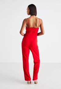 Rød jumpsuit med justerbare spaghetti-stropper, tettsittende liv og løse bein. Glatt stoff med en lett glans, med baklommer.