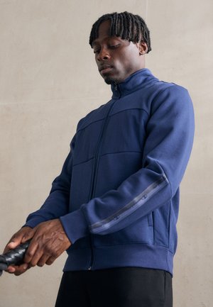 Chaqueta azul marino con cremallera y cuello alto, paneles texturizados y detalles reflectantes en las mangas, con un diseño ajustado y bolsillos.