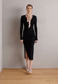 Robe noire à manches longues avec une silhouette ajustée et une fente sur le côté. Associée à une écharpe crème avec des rayures brunes et des talons argentés.