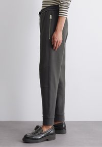 Pantalones jogger grises con cintura ajustable con cordón, bolsillos laterales con cremallera y piernas ajustadas, combinados con zapatos negros sin cordones. Textura suave.