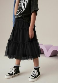 Jupe en tulle noire avec des volants superposés, portée avec un t-shirt graphique noir et des baskets montantes noires, mettant en valeur la texture et le design en couches.