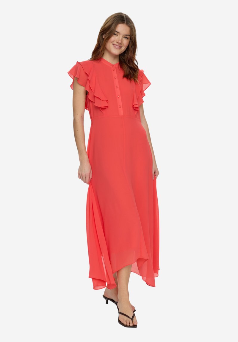 Jeune femme portant une robe maxi corail à manches volantées et devant boutonné, souriante et tenant l'ourlet de la robe, portant des sandales noires à talons.