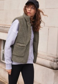 Vesta fără mâneci de culoare verde măsliniu, dintr-un material fleece texturat, cu un buzunar tip patch, purtată peste un hanorac gri și colanți negri. Pe cap este purtată o șapcă neagră.