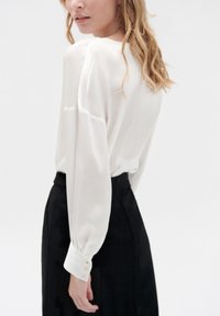 Weiße Bluse mit langen Ärmeln, weicher Stoff, mit Knopfakzenten an den Manschetten, kombiniert mit einem schwarzen Rock, einfaches Design und minimalistischer Stil.