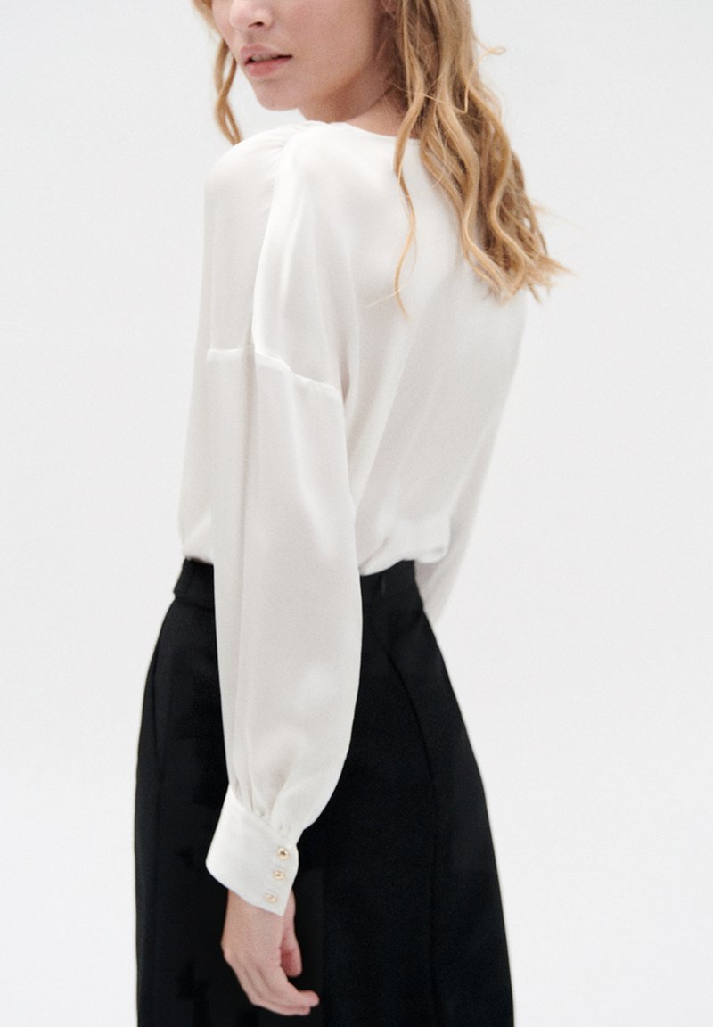 Weiße Bluse mit langen Ärmeln, weicher Stoff, mit Knopfakzenten an den Manschetten, kombiniert mit einem schwarzen Rock, einfaches Design und minimalistischer Stil.