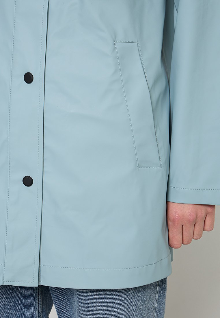 Chaqueta azul claro con botones a presión negros, bolsillo frontal en ángulo, llevada por una persona cuya mano es visible al lado sobre unos vaqueros azul.