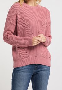 Růžový pletený svetr s ribováním, geometrickými vzory a malými akcentovými otvory poblíž ramen. Kombinovaný s modrými džíny.