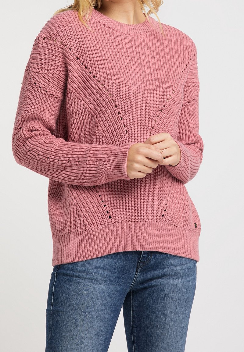 Růžový pletený svetr s ribováním, geometrickými vzory a malými akcentovými otvory poblíž ramen. Kombinovaný s modrými džíny.