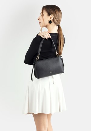 Donna con capelli lunghi, indossa un top nero e una gonna bianca, con una borsa a tracolla in pelle nera sulla spalla, vista di profilo su sfondo bianco.