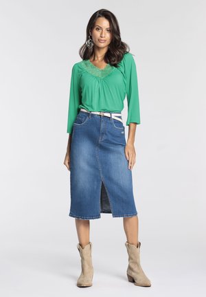 Blouse verte avec des détails en dentelle au niveau du col, associée à une jupe en denim avec une fente à l'avant. Des bottines beiges à la cheville complètent la tenue.