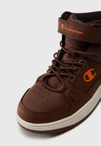 Zapatilla alta marrón hecha de material sintético, con el logotipo de Champion en naranja, punta redondeada, detalles perforados y tobillo acolchado.