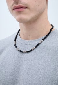 Fekete gyöngy nyaklánc, ezüst és bronz elemek váltakozásával, egy szürke pulóver fölött viselve. A nyakkivágás látható.