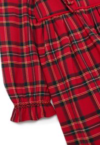 Roter Karostoff mit schwarzen, weißen und gelben Streifen, der an einem Kleidungsstück mit gerafftem Ärmel, elastischem Bündchen und Rüschendetail versehen ist.