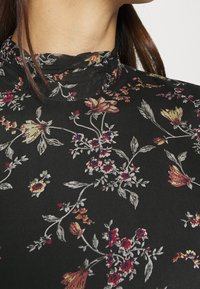 Blouse florale noire avec un col montant, un tissu délicat, ornée de motifs floraux multicolores en rouge, jaune et bleu sur un fond sombre.