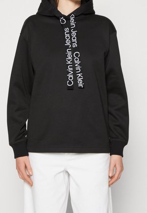 Osoba ubrana w czarną bluzę z kapturem Calvin Klein z logo na sznurkach i białe spodnie, stojąca na tle jasnego, jednolitego tła.
