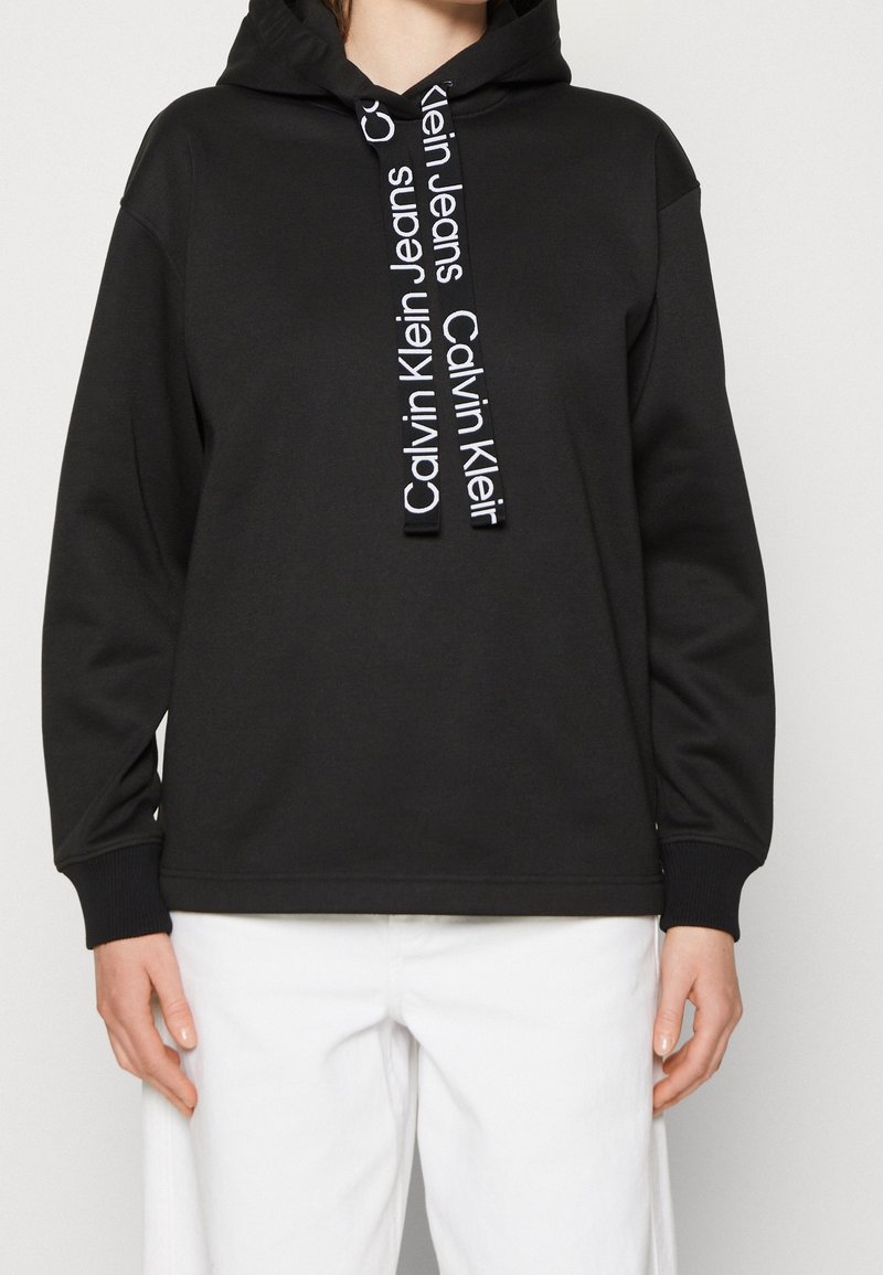 Personne portant un sweat à capuche noir Calvin Klein avec logo sur les cordons et un pantalon blanc, debout devant un fond uni clair.