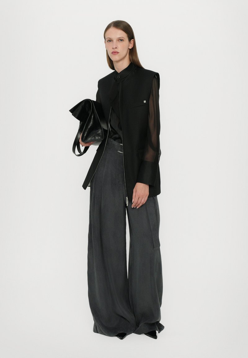 Black sleeveless blazer over a black shirt, paired with wide gray trousers. The outfit includes a black handbag with smooth texture.