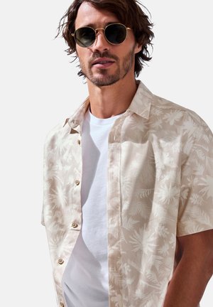 Kurzarm-Hemd in hellbeige mit weißen Blattmustern, das eine durchgehende Knopfleiste und eine weiche, glatte Textur aufweist, über einem weißen T-Shirt getragen.