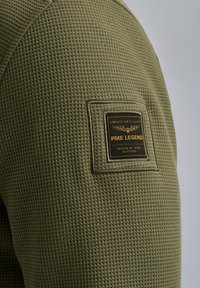 Olijfgroene longsleeve shirt van textuur met wafelstructuur. Heeft een vierkante zwart-gouden merkpatch op de bovenarm.