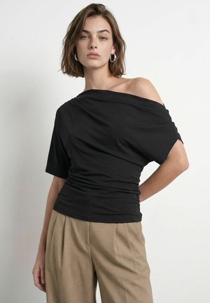 Jeune femme aux cheveux bruns courts portant un top noir épaule dénudée et un pantalon beige à jambes larges sur un fond blanc uni.