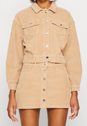 Femme portant une veste boutonnée en velours côtelé beige et une mini-jupe boutonnée taille haute assortie, les bras détendus le long du corps.