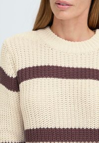 Kremfarget strikket genser med maroon horisontale striper. Har rund hals og teksturert stoff. Lett og uformell design.