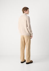 Beige kötött pulóver magas gallérral, világos bézs nadrággal párosítva. A pulóver bordázott textúrával és laza fit-tel rendelkezik.