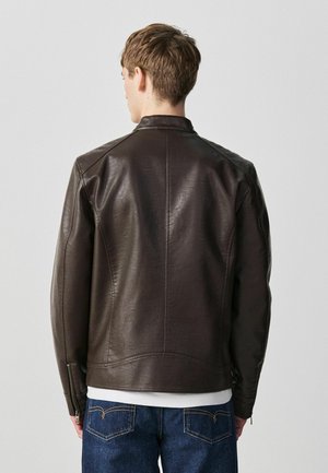 Mann, der eine dunkelbraune Lederjacke und blaue Jeans trägt, von hinten vor einem einfarbigen hellgrauen Hintergrund gezeigt.