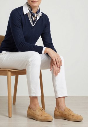 Kvinde sidder på træstol iført marineblå sweater, hvide bukser, mønstret halstørklæde og beige ruskindssnøresko på lys gulv.
