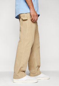 Beżowe bawełniane spodnie o luźnym kroju, z bocznymi kieszeniami, przeszyciami i wywiniętymi mankietami, zestawione z białymi trampkami.