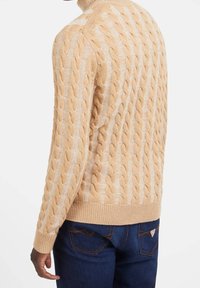 Béžový pletený svetr s copánkovým vzorem a vysokým límcem; žebrované manžety a spodní lem; nošený s tmavě modrými džínami.
