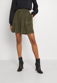 Falda de ante sintético en color verde oliva con un diseño en A, dos bolsillos delanteros y una cintura ajustada. Combinada con botas negras hasta el tobillo.