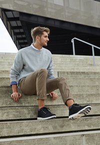Svarta mocka anklarboots med vita sulor, grå sweatshirt och bruna uppvikta byxor. Modellen sitter på betongtrappor i en modern utomhusmiljö.