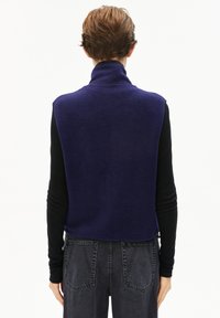 Marineblaue Fleece-Weste mit hohem Kragen, Rückenschlitz und ohne Ärmel, getragen über einem schwarzen Langarmshirt, kombiniert mit dunklen Jeans.
