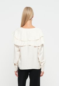 Crème-kleurige blouse met gelaagd ruchesontwerp op de rug, lange mouwen en met knopen sluitende manchetten, gemaakt van een lichtgewicht, textuurstof.