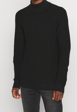 Schwarzer Strickpullover mit hohem, geripptem Kragen, Raglanärmeln und strukturiertem Stoff. Mit einem kleinen Logostickerei am unteren Saum.