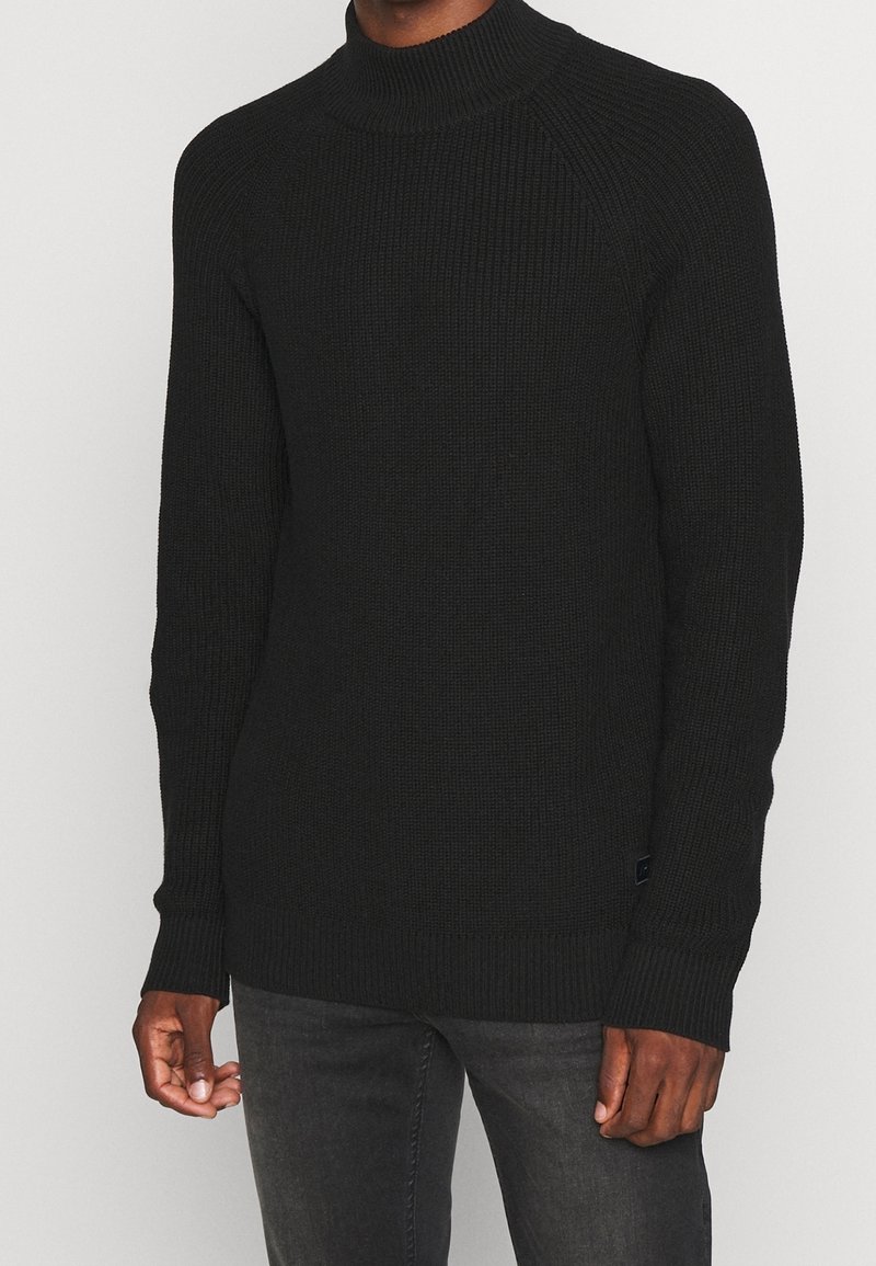 Svart stickad tröja med hög, ribbad krage, raglanknappar och textilstruktur. Har en liten logotagg vid nederkanten.