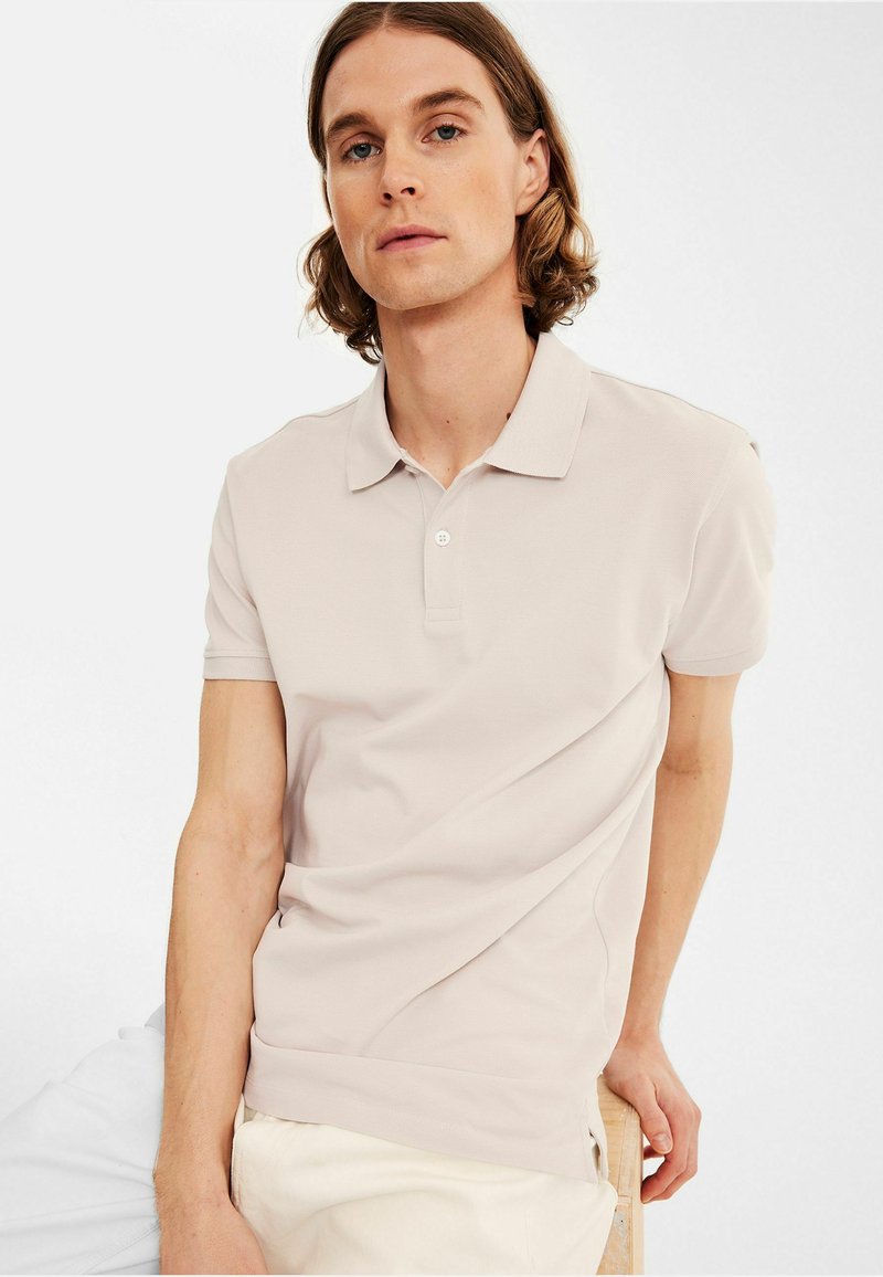 Ljust beige pikétröja med krage, korta ärmar och en strömlinjeformad design. Har ett subtilt strukturerat tyg och en två-buttons knappslå.