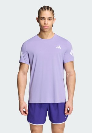Giovane uomo con i capelli intrecciati che indossa una maglietta Adidas viola chiaro e pantaloncini viola scuro, in piedi davanti a uno sfondo semplice.