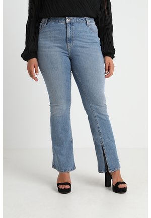 Frau trägt hellblaue High-Waist-Jeans mit vorderen Schlitzen, kombiniert mit schwarzen Plateau-Open-Toe-Heels und einem schwarzen, strukturierten Langarmtop.