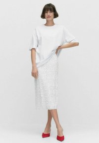 Haut oversize blanc, jupe midi blanche à fleurs texturées et chaussures rouges à bout pointu. Matériaux élégants, design moderne et détails uniques.