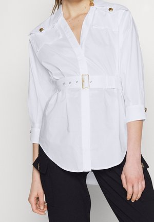 Kobieta ubrana w białą koszulę z paskiem i złotymi guzikami na ramionach i mankietach, w połączeniu z czarnymi spodniami bojówkami z kieszeniami zapinanymi na guziki.