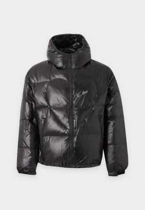 Homme portant une doudoune noire brillante, une chemise rose, un pantalon large beige et des chaussures noires, debout les mains dans les poches de la veste.