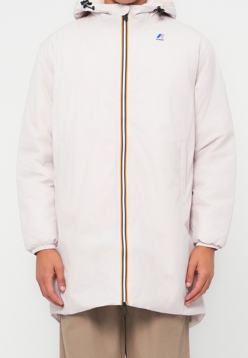 Personne portant une veste d’hiver à capuche beige clair avec une fermeture éclair multicolore à l’avant et un petit logo sur la poitrine.