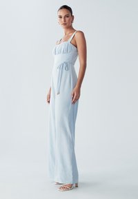 Svetlo modra kombinezona z oprijetim telesom, širokimi hlačnicami in nastavljivim pasom. Ima tanke naramnice in subtilno teksturo. Kombinirano z belimi čeveljci.