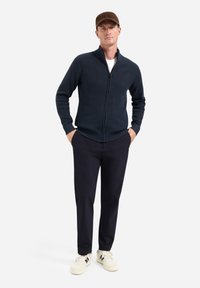 Marineblauwe zip-up trui met een textuurpatroon, gecombineerd met donkere broeken en beige sneakers. Toegankelijke pasvorm en functionele ontwerpdetaillering.