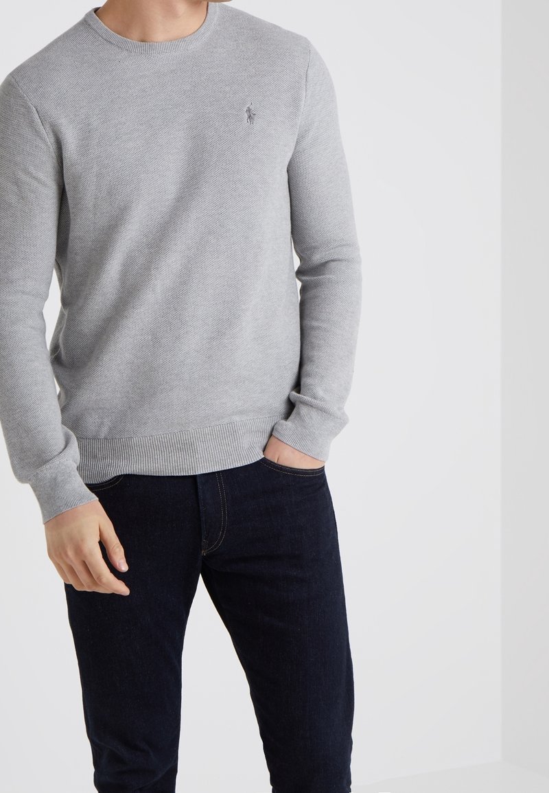 Homme portant un pull en maille gris clair et un jean bleu foncé, une main dans la poche, sur fond blanc uni.