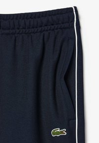 Pantalon à taille élastique bleu marine avec poche latérale et détail de rayure blanche, arborant un petit logo crocodile vert près de la poche.